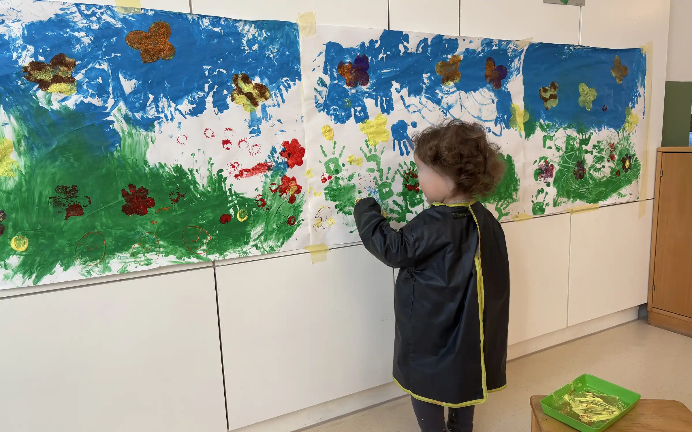 Ein Kind steht an einer Wand und malt mit Hilfe seiner Hände eine Wiese, einen Himmel und Schmetterlinge.