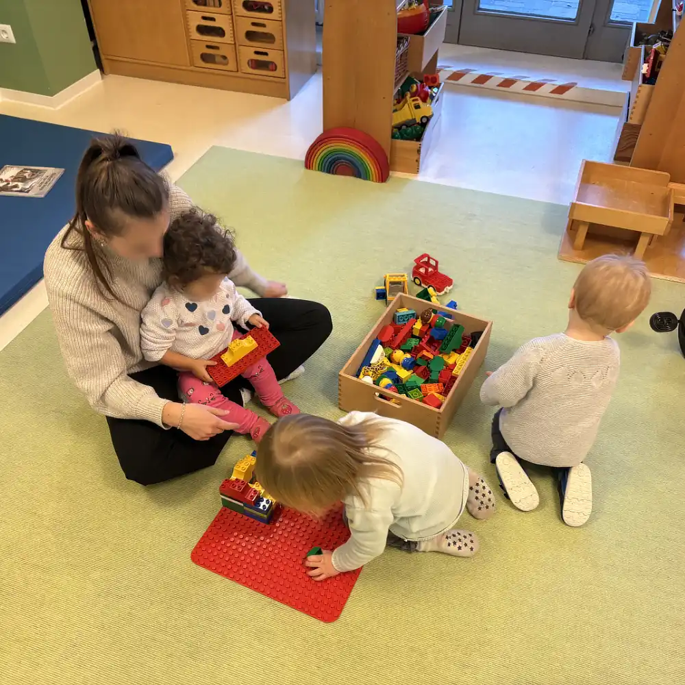 Kinder spielen mit Lego und Erzieherin liest einem Kind ein Buch vor.