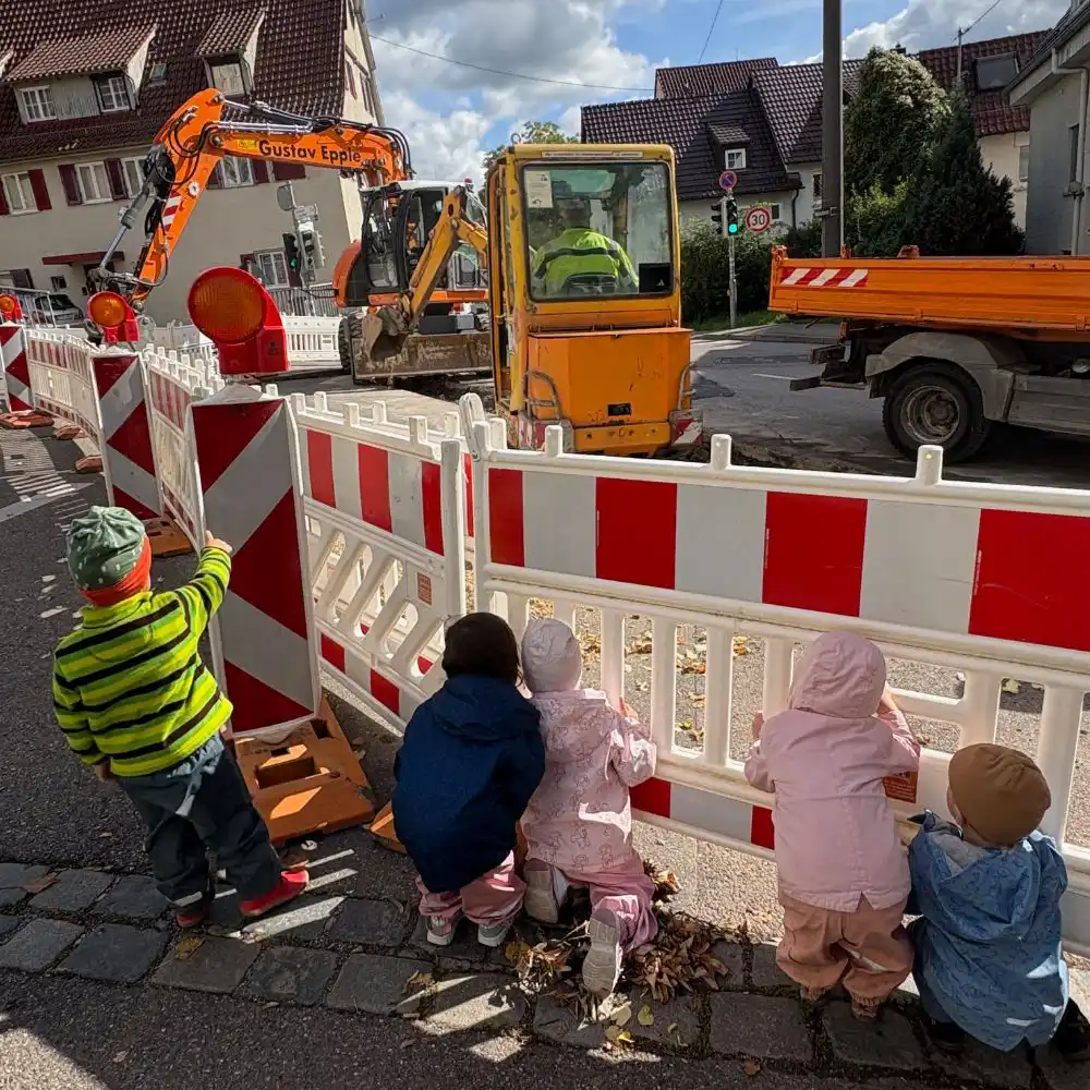 Kinder entdecken beim Spaziergang eine Baustelle und betrachten gespannt den Bagger durch den Bauzaun.
